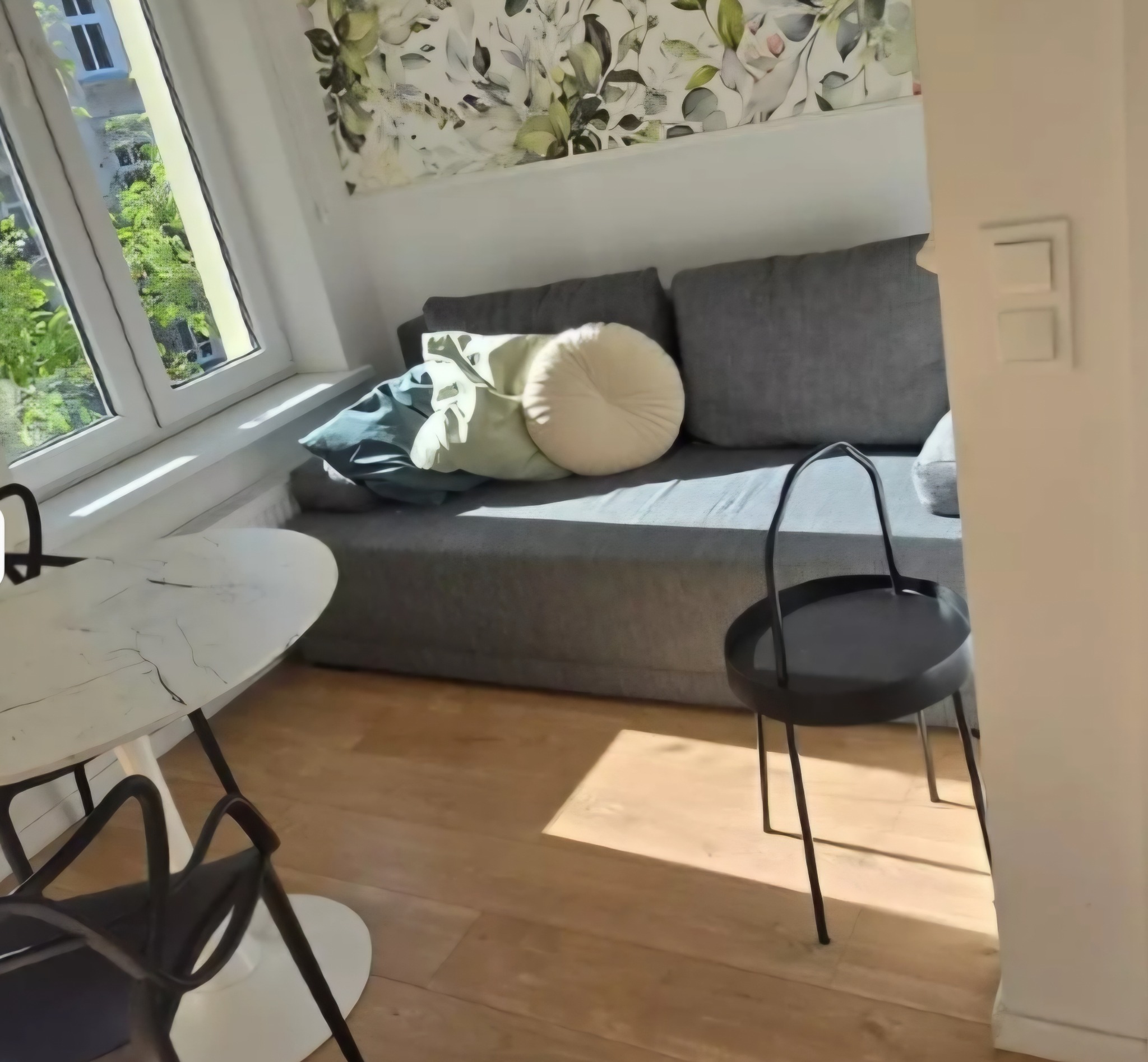 Jasne wnętrze pokoju z drewnianą podłogą, szarą sofą z poduszkami, okrągłym stolikiem i krzesłem. Widok z okna na zieleń. Tapeta w kwiaty.