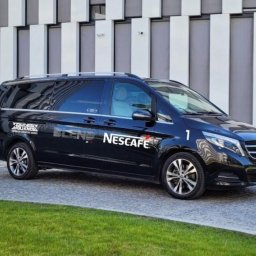 DPIVP SPÓŁKA Z OGRANICZONĄ ODPOWIEDZIALNOŚCIĄ - Czarny bus Mercedes z reklamą Nescafe Blend, zaparkowany przed nowoczesnym budynkiem z pionowymi panelami. Widoczne logo i numer '1' na karoserii, koła aluminiowe.