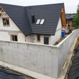 Dom murowany w trakcie budowy z widocznymi fundamentami, szarą dachówką, oknami z drewnianymi ramami, otoczony betonowym murem oporowym i folią zabezpieczającą.