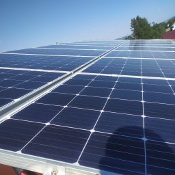 VOLTOM Tomasz Kondej - Błękitne panele słoneczne na dachu domu w słoneczny dzień. Widoczne góry i drzewa w tle. Czyste niebo i cień na panelach.