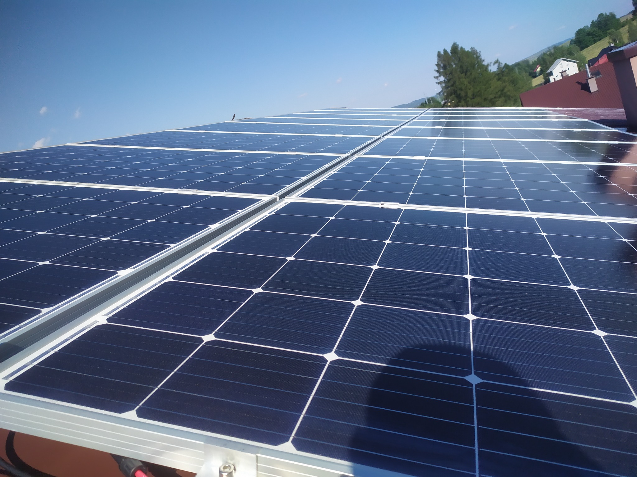 Błękitne panele słoneczne na dachu domu w słoneczny dzień. Widoczne góry i drzewa w tle. Czyste niebo i cień na panelach.