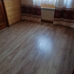 Karol Cież - Jasne panele podłogowe w pokoju z drewnianą boazerią i zasłonami w paski. Widoczny fragment grzejnika pod oknem. Ujęcie z niskiego kąta.