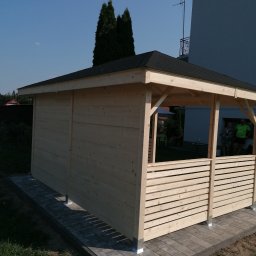 Karol Cież - Nowo postawiona drewniana wiata ogrodowa z jasnego drewna, z czarnym dachem i ażurowymi panelami bocznymi, osadzona na szarych płytach chodnikowych.