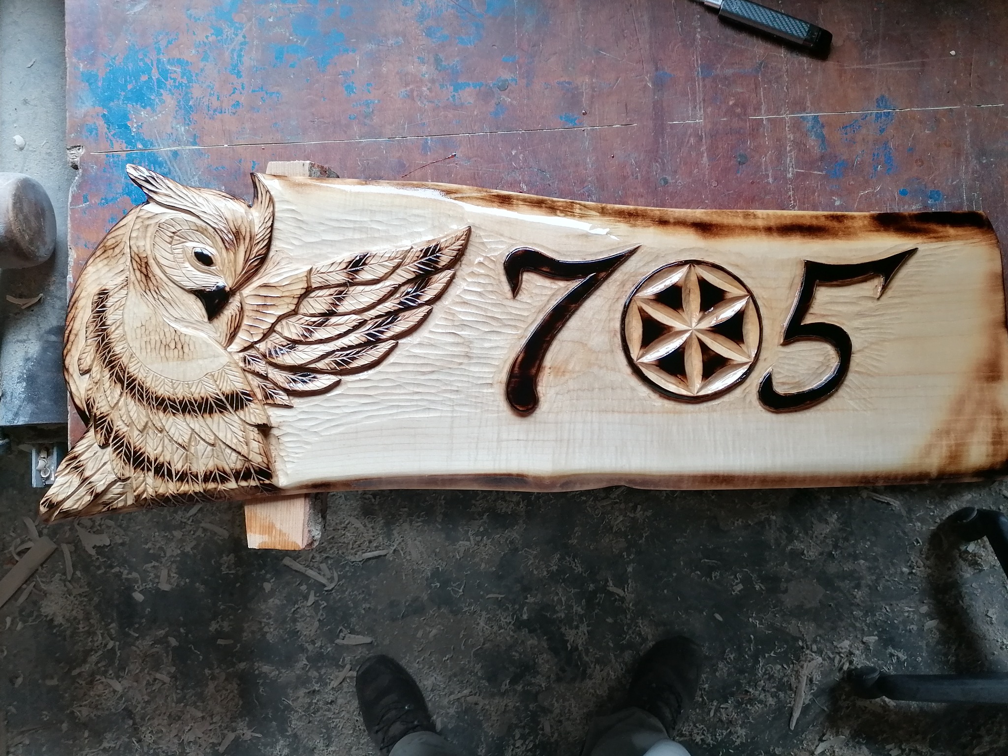Ręcznie rzeźbiony, drewniany szyld z numerem '705' i motywem sowy, leżący na warsztatowym stole. Widoczne narzędzie i wióry, podkreślające unikatowy charakter.