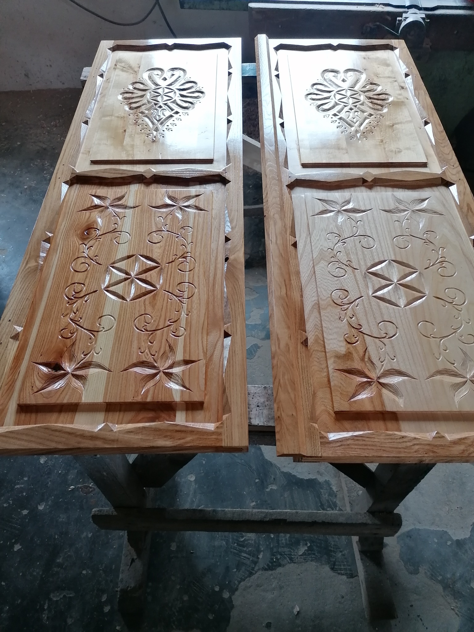 Dwie drewniane płyty z rzeźbieniami kwiatowymi i geometrycznymi, leżące na drewnianym stole. Widoczne detale rzeźb i naturalne usłojenie drewna.
