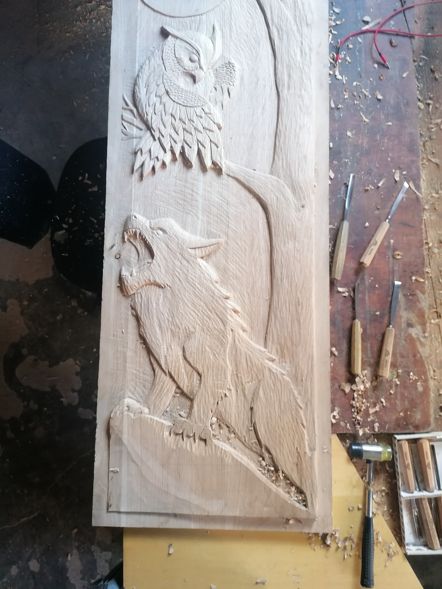 Ręcznie rzeźbiona w drewnie płaskorzeźba przedstawiająca wyjącego wilka pod sową, otoczona dłutami i wiórami, ukazująca precyzję i kunszt artystyczny.