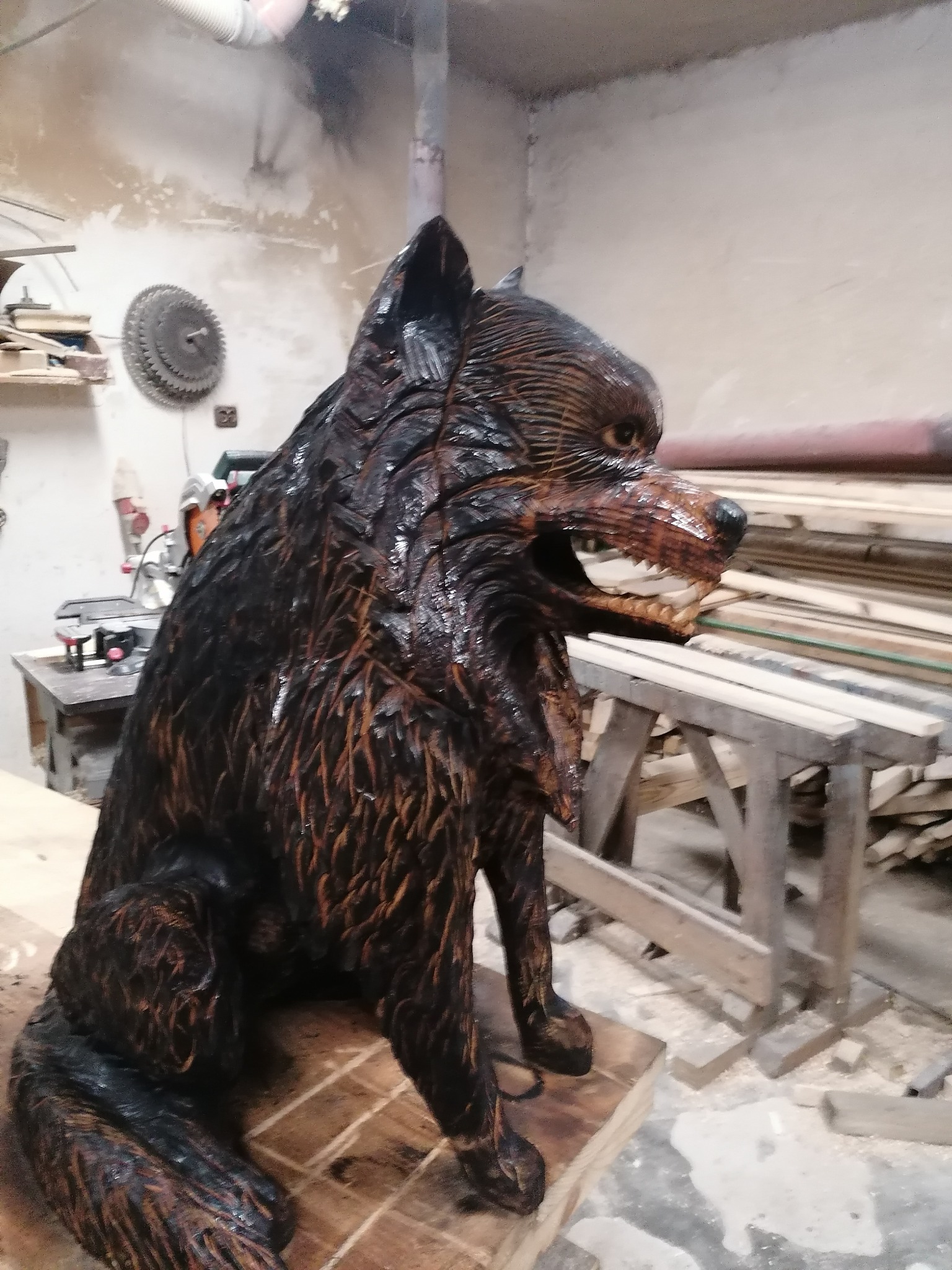 Ręcznie rzeźbiona, lakierowana figura wilka z drewna, prezentowana w warsztacie w Kasince Małej. Widoczne detale rzeźby i narzędzia stolarskie w tle.