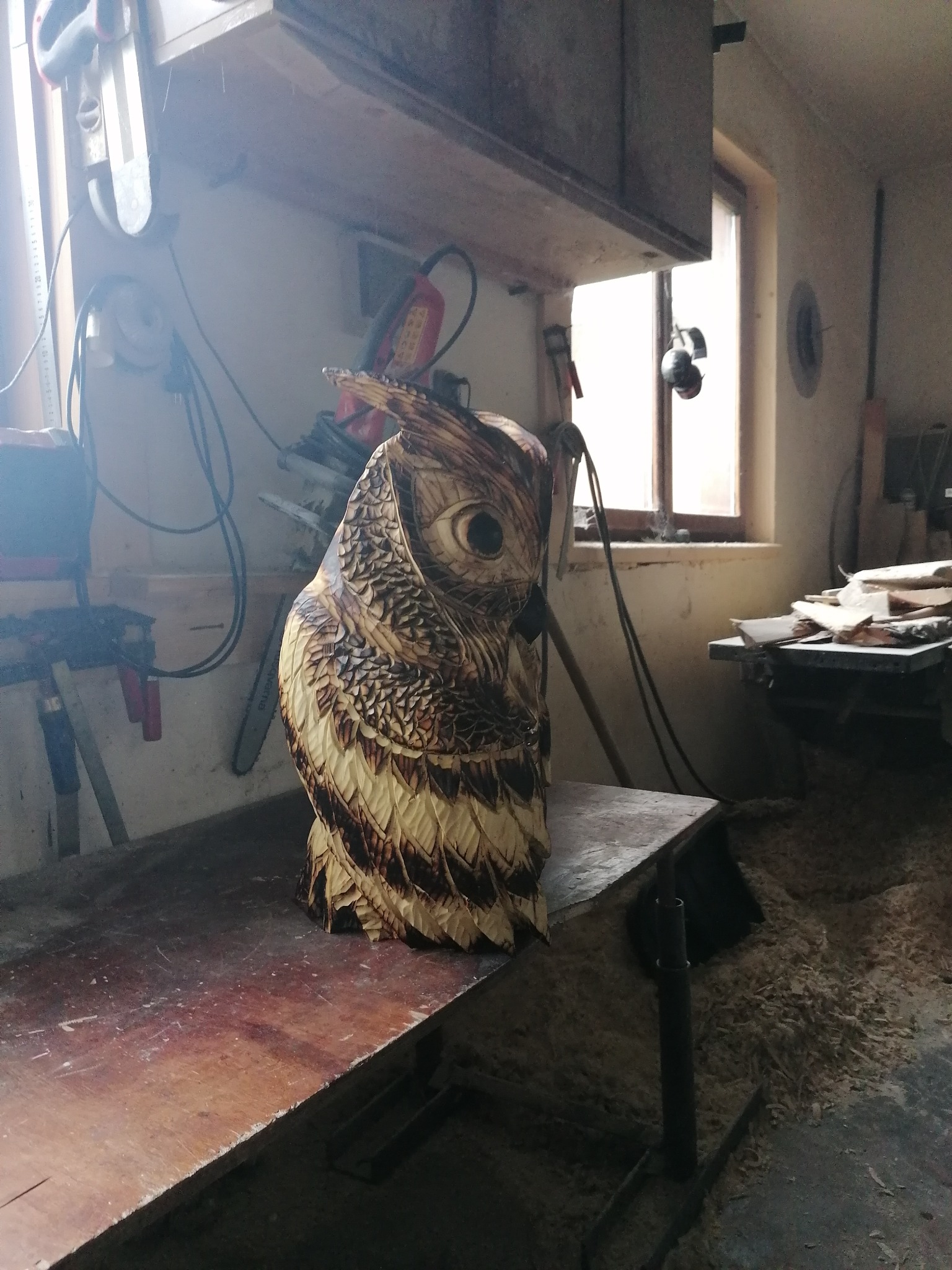 Ręcznie rzeźbiona, drewniana figura sowy z detalami piór, umieszczona w warsztacie stolarskim z narzędziami i oknem w tle. Unikalne rzemiosło artystyczne.