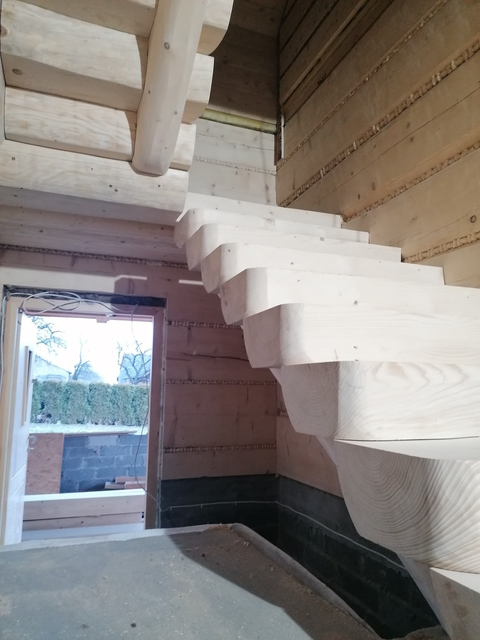 Drewniane schody w surowym wnętrzu z bali, widok z dołu. Jasne drewno, widoczne słoje. W tle otwarte drzwi z widokiem na zewnątrz.