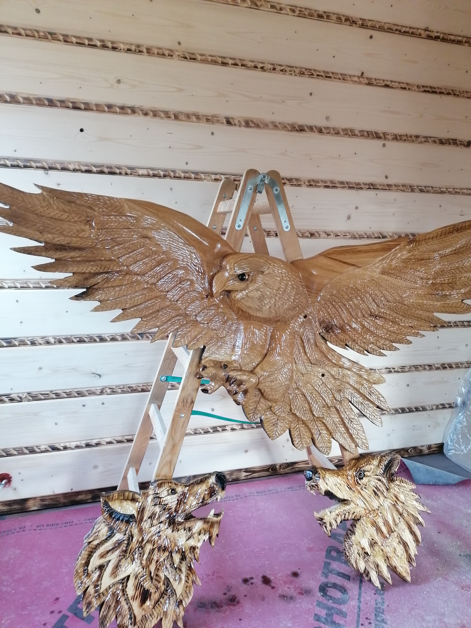 Ręcznie rzeźbiony, drewniany orzeł z rozpostartymi skrzydłami na tle drewnianej ściany, poniżej dwie rzeźbione głowy wilków. Widoczna drabina.