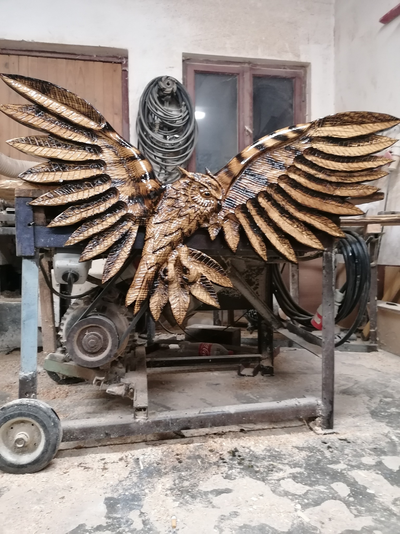 Ręcznie rzeźbiona, drewniana figura sowy z rozpostartymi skrzydłami, umieszczona na metalowym stole warsztatowym w pracowni. Widoczne detale rzeźby i faktura drewna.