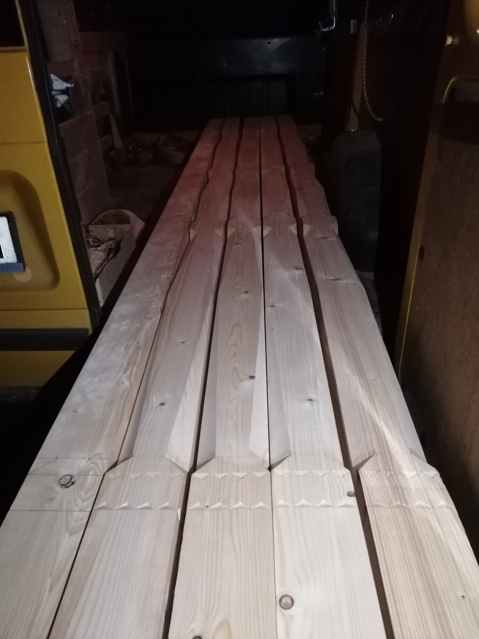 Przygotowane deski z wyżłobieniami, prawdopodobnie do altany, ułożone wewnątrz pojazdu dostawczego. Widoczne ślady ołówka i sęki w drewnie.