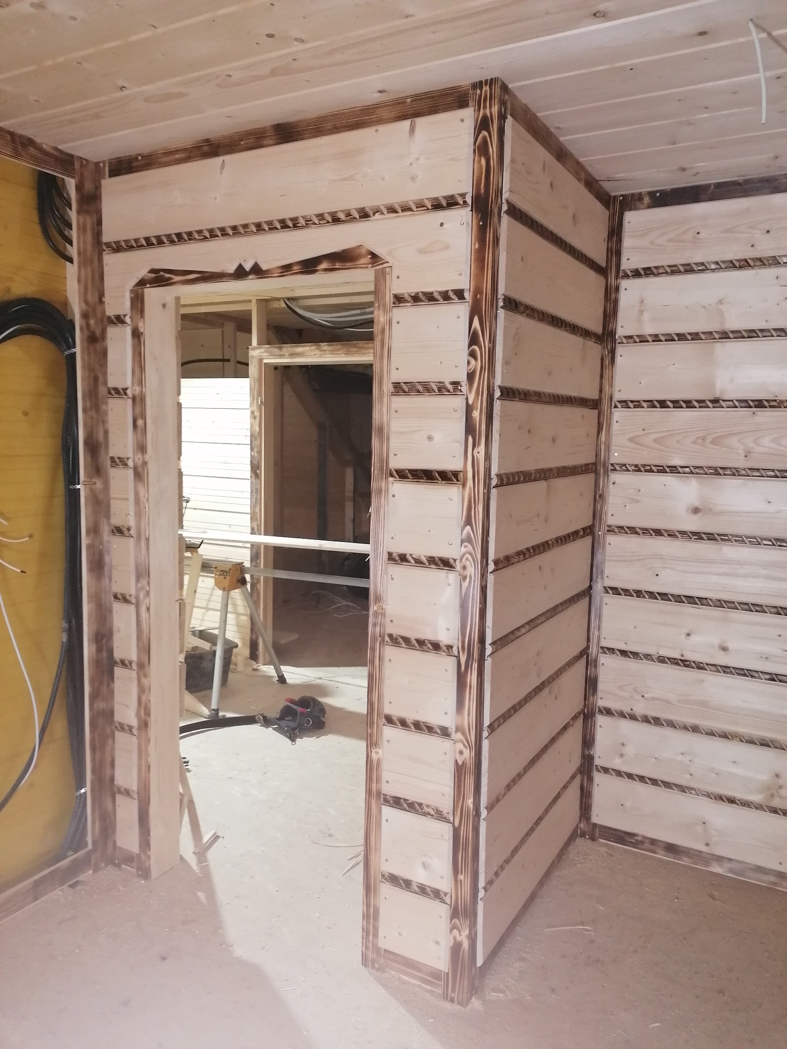 Wnętrze w trakcie wykańczania boazerią z jasnego drewna z ciemnymi akcentami, widoczne narzędzia i elementy konstrukcyjne. Prace wykończeniowe w pomieszczeniu.