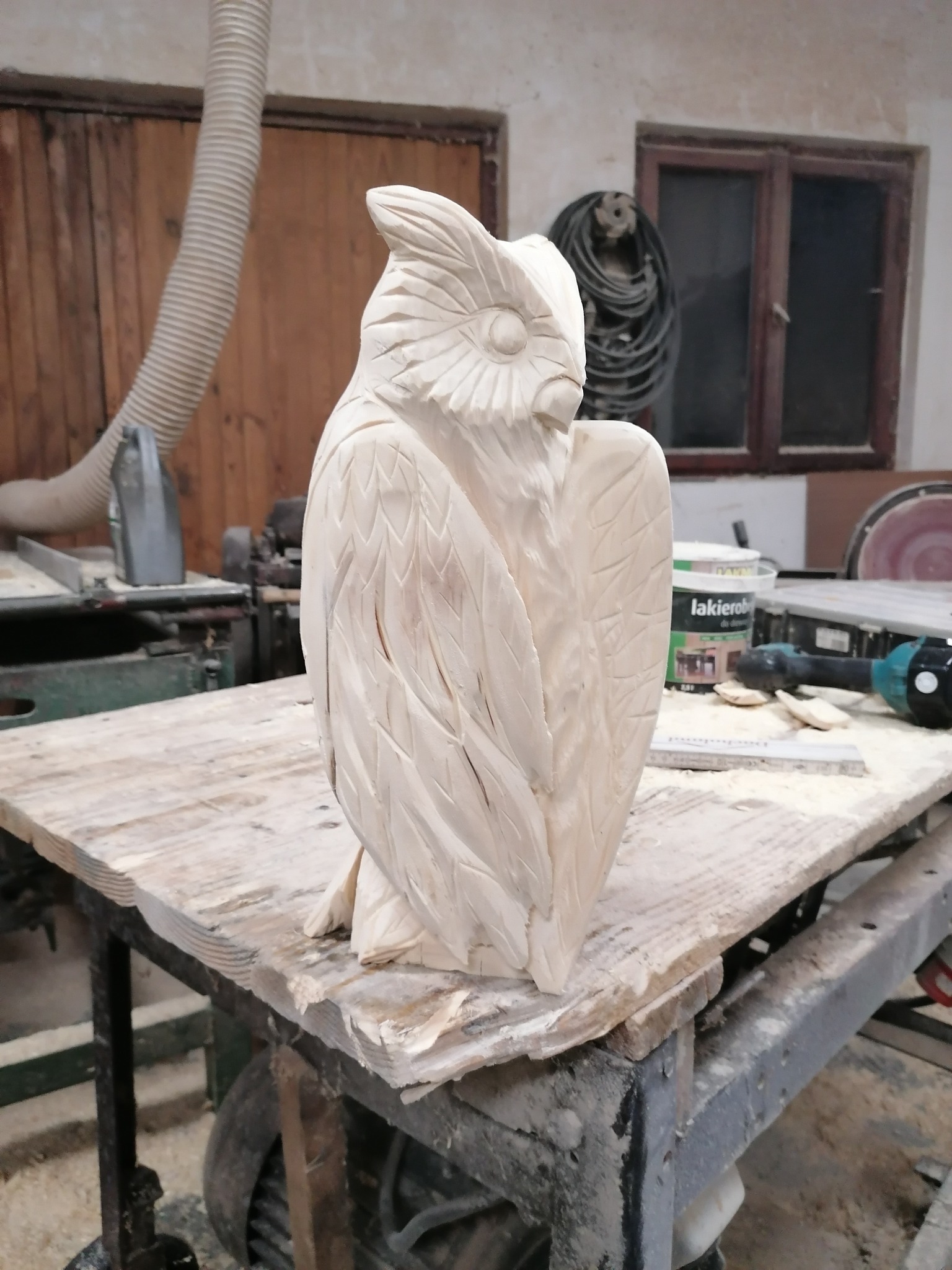 Ręcznie rzeźbiona, surowa figura sowy z drewna na warsztatowym stole, z widocznymi narzędziami i puszką lakierobejcy w tle. Detaliczna praca snycerska.