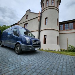 WenaBud - Niebieski bus dostawczy Volkswagen Crafter zaparkowany na kostce brukowej przed budynkiem z wieżą w stylu zamkowym, pochmurne niebo w tle.