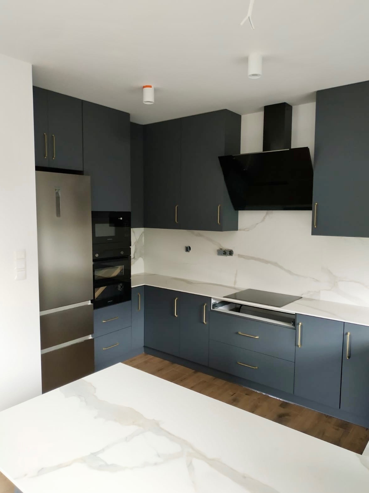 Nowoczesna kuchnia w odcieniach szarości z marmurowym blatem i złotymi uchwytami. Widoczny sprzęt AGD w zabudowie i minimalistyczny okap.