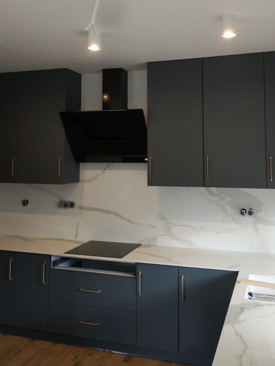 Nowoczesna, grafitowa kuchnia z białym blatem imitującym marmur i złotymi uchwytami. Widoczna płyta indukcyjna i minimalistyczny okap. Oświetlenie punktowe.