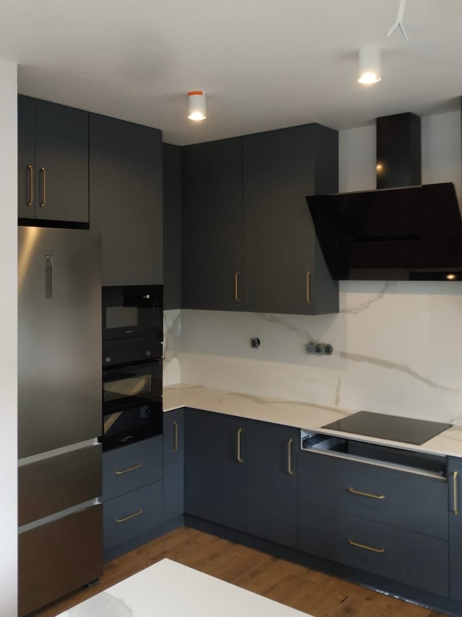 Nowoczesna kuchnia w odcieniach szarości z marmurowym blatem i złotymi uchwytami, oświetlona punktowymi lampami sufitowymi. Widoczny sprzęt AGD w zabudowie.