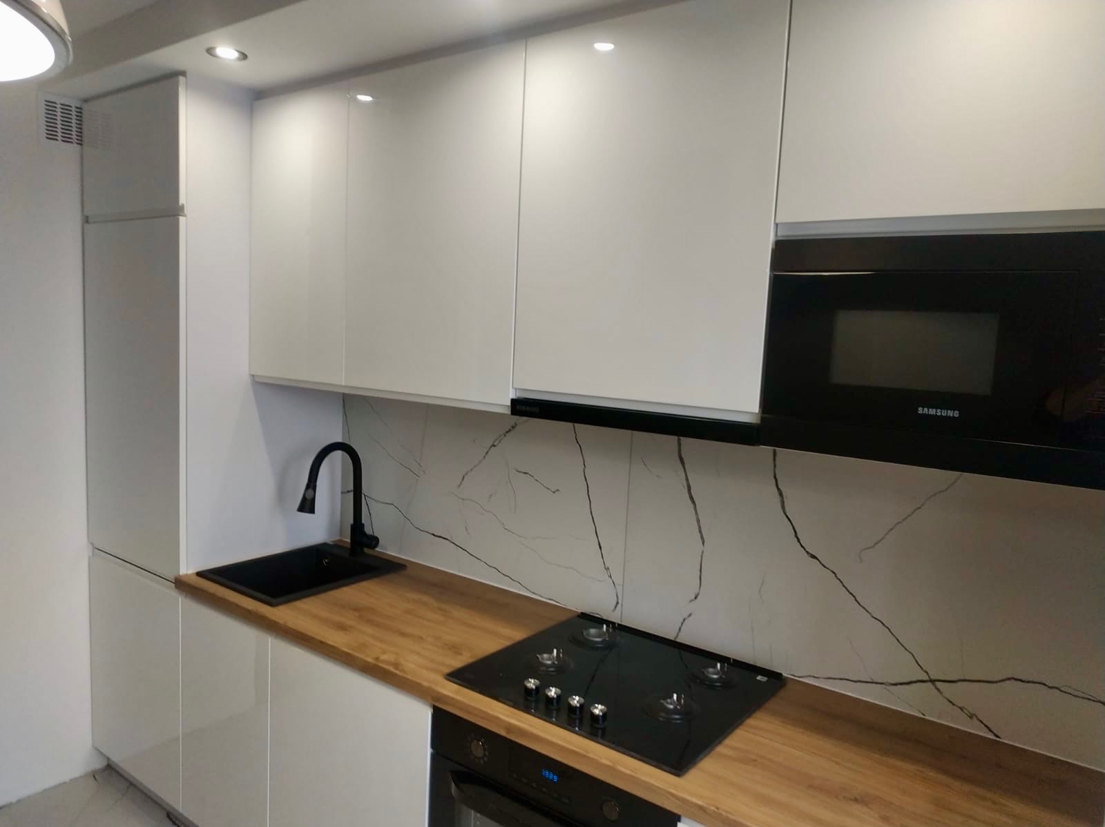 Biała kuchnia z drewnianym blatem, czarnym zlewem i płytą grzewczą. Marmurowy wzór na ścianie. Nowoczesne oświetlenie punktowe.