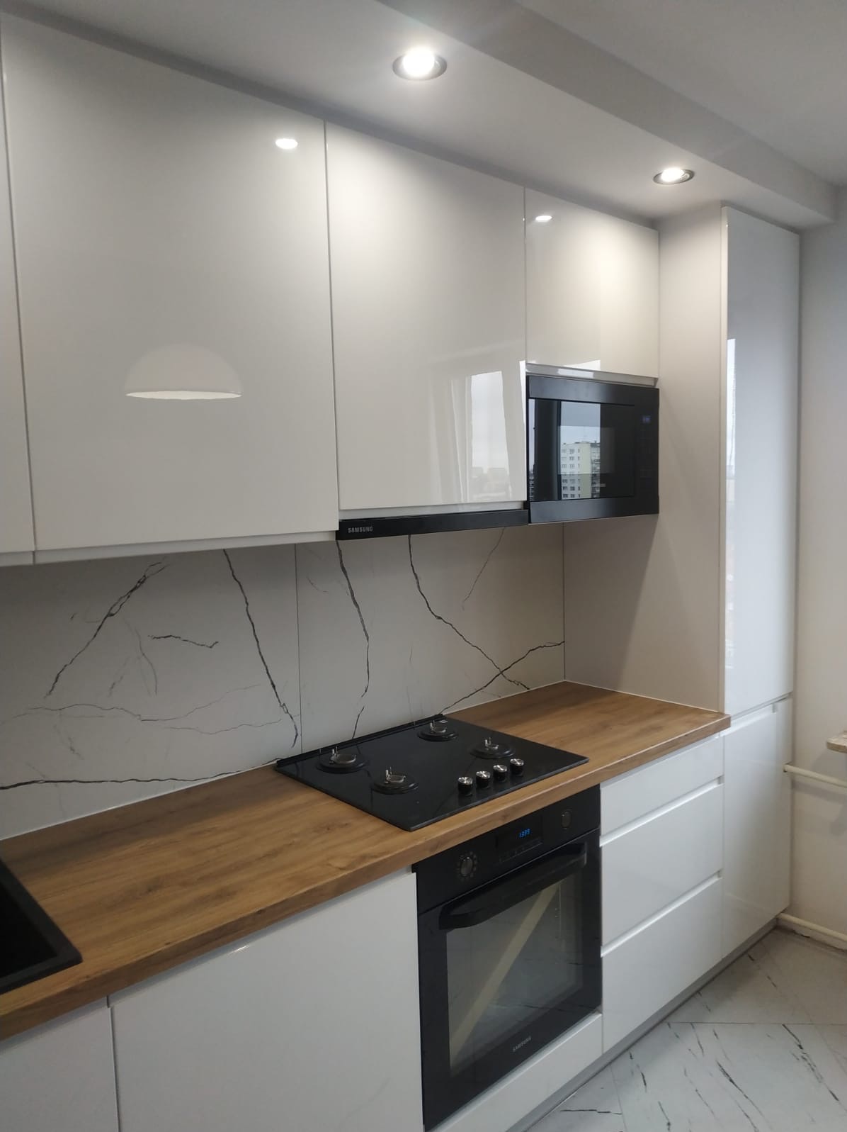 Biała, nowoczesna kuchnia z drewnianym blatem i marmurowym backsplashiem. Widoczna płyta grzewcza i piekarnik. Minimalistyczny design z błyszczącymi frontami szafek.