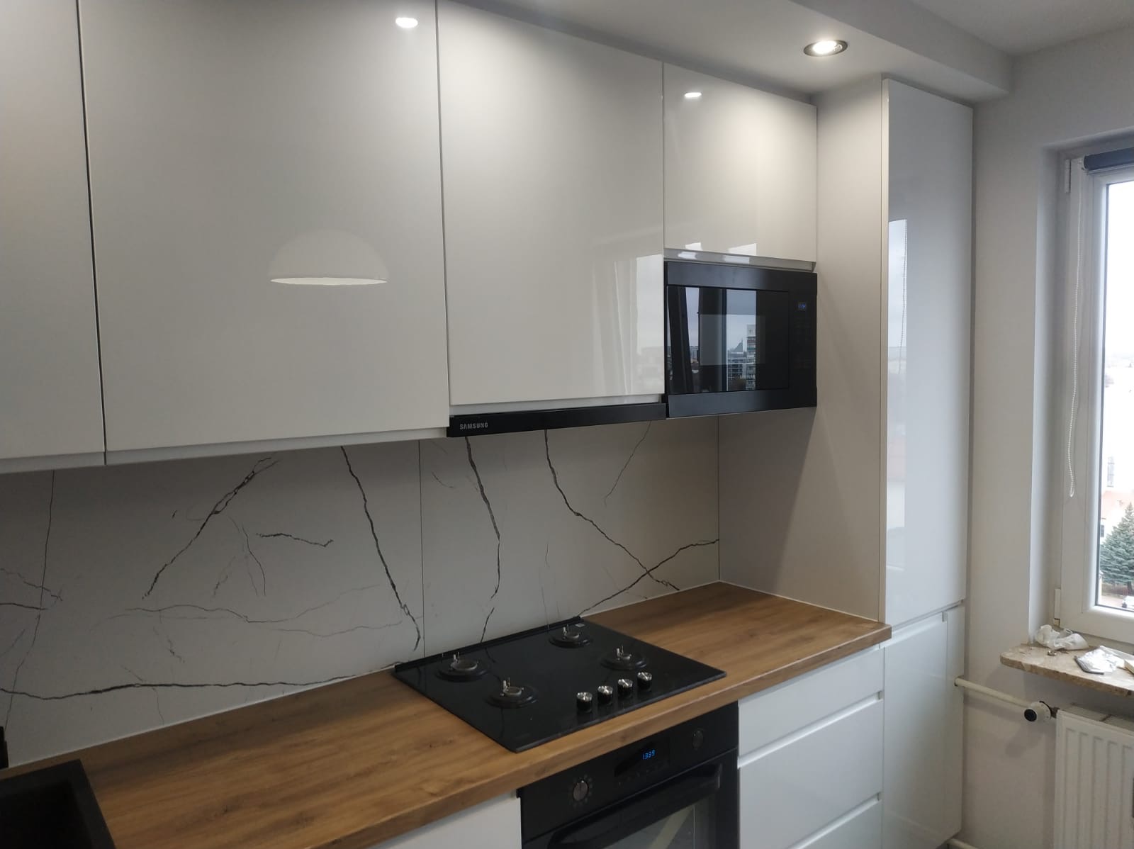 Biała kuchnia z połyskiem, drewniany blat i marmurowy backsplash. Nowoczesny design z piekarnikiem i płytą grzewczą. Minimalistyczne oświetlenie punktowe.