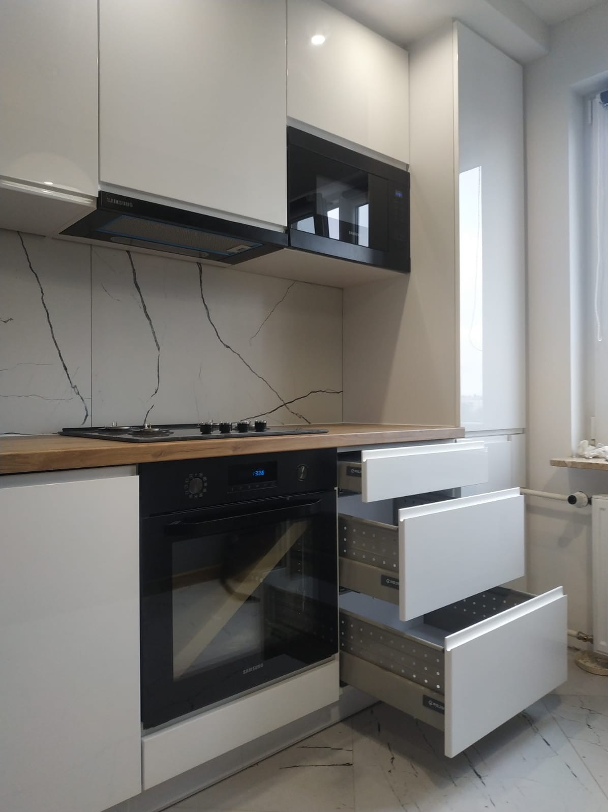 Biała kuchnia z drewnianym blatem i otwartymi szufladami. Płytki imitujące marmur na ścianie. Nowoczesny design z piekarnikiem Samsung i czarną mikrofalówką.