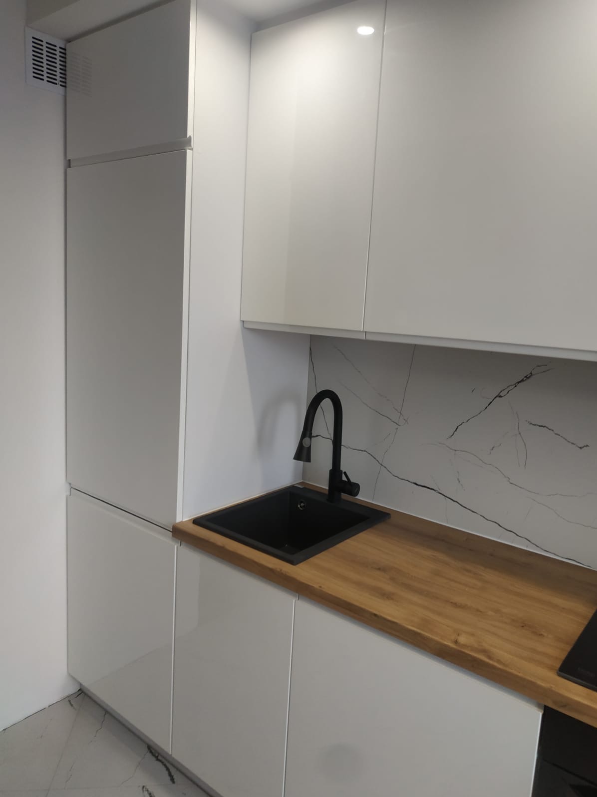 Biała, nowoczesna kuchnia z drewnianym blatem i czarnym zlewem. Minimalistyczny design, gładkie fronty szafek, marmurowe wykończenie ściany nad blatem. Oświetlenie punktowe.