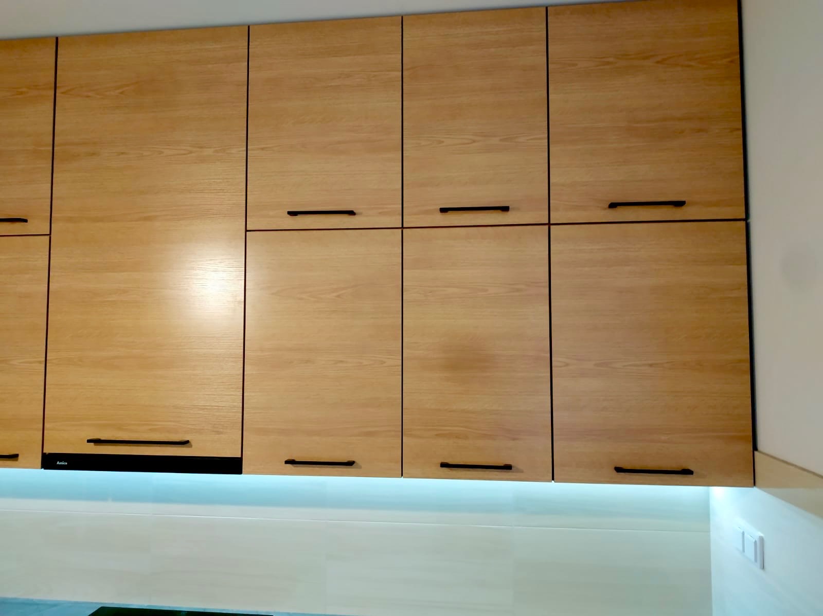 Drewniane szafki kuchenne górne z czarnymi, minimalistycznymi uchwytami i podświetleniem LED. Fronty z wyraźnym usłojeniem. Widoczny fragment blatu.