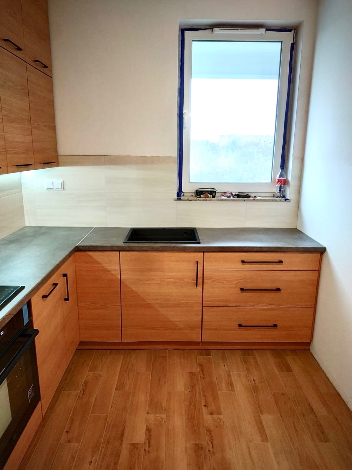 Jasna kuchnia z drewnianymi frontami i ciemnym blatem, widok na okno z akcesoriami montażowymi. Uwagę przykuwają minimalistyczne, czarne uchwyty.