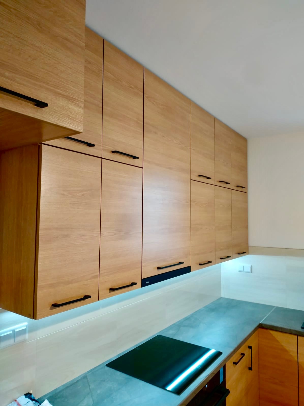 Jasne, drewniane szafki kuchenne z czarnymi uchwytami, oświetlenie LED pod szafkami, szary blat i płyta indukcyjna. Ujęcie z dołu, podkreślające wysokość zabudowy.