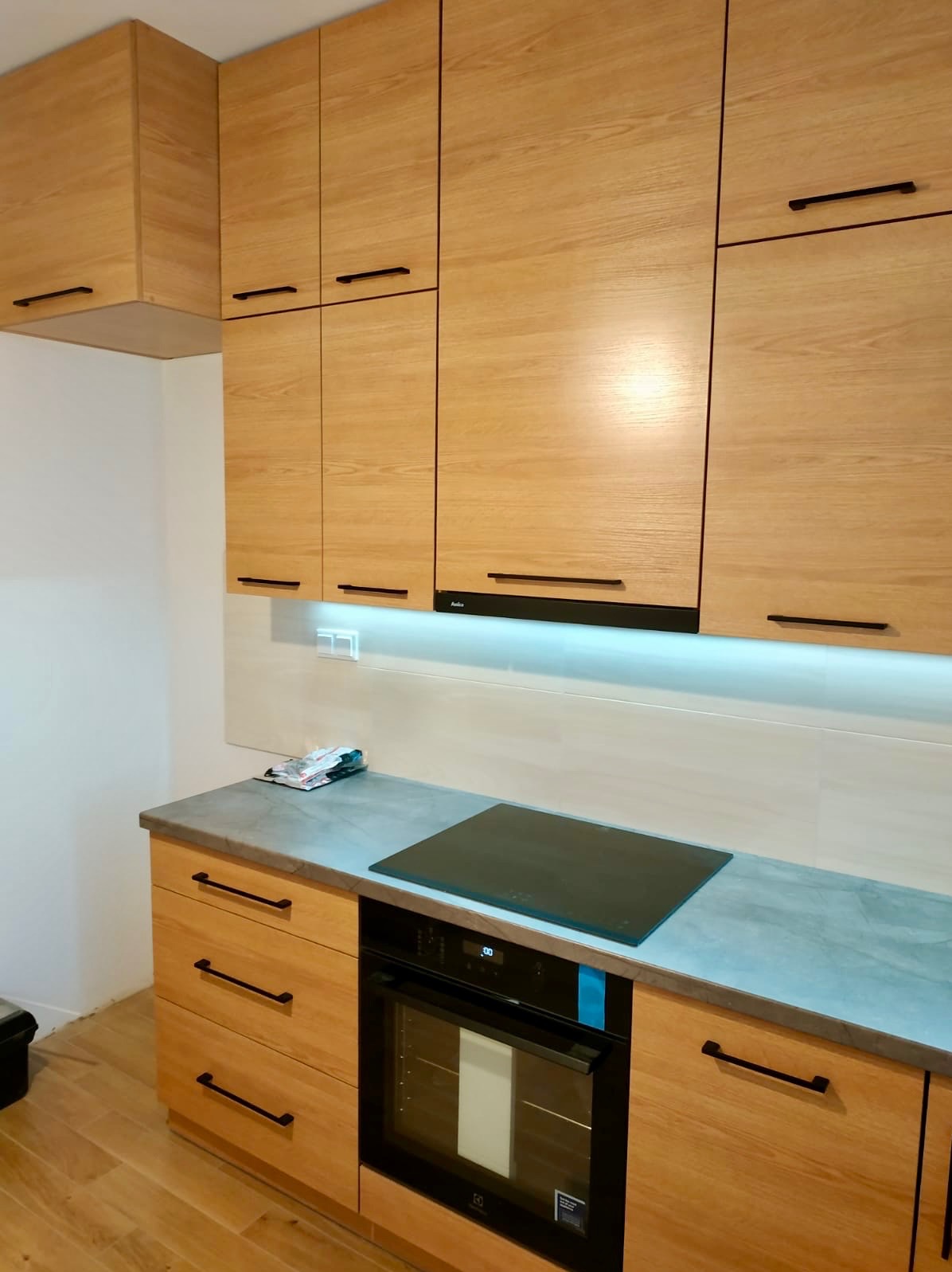 Jasna, drewniana zabudowa kuchenna z czarnymi uchwytami, szary blat, płyta indukcyjna i piekarnik. Ujęcie z perspektywy podłogi, widoczne fragmenty podłogi i ściany.