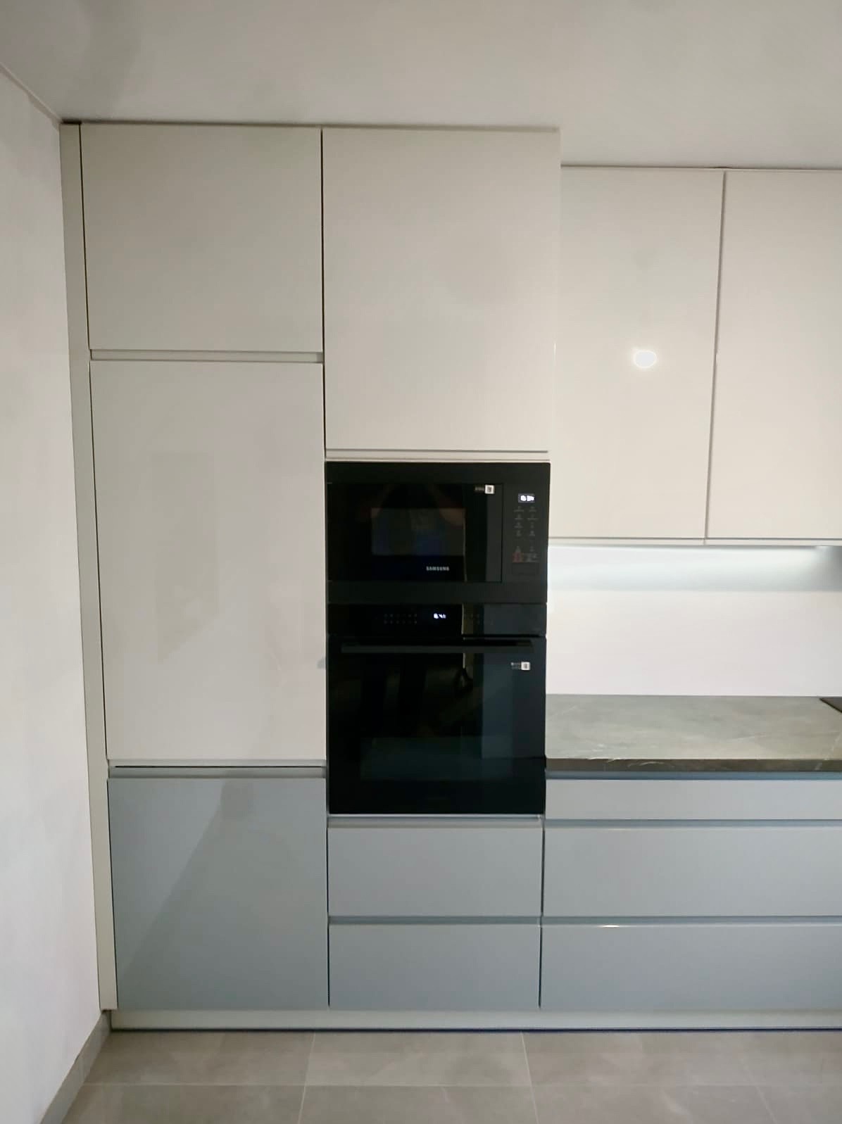 Nowoczesna zabudowa kuchenna z piekarnikiem i mikrofalówką Samsung w kolorach bieli i szarości. Minimalistyczny design, gładkie fronty, brak uchwytów.