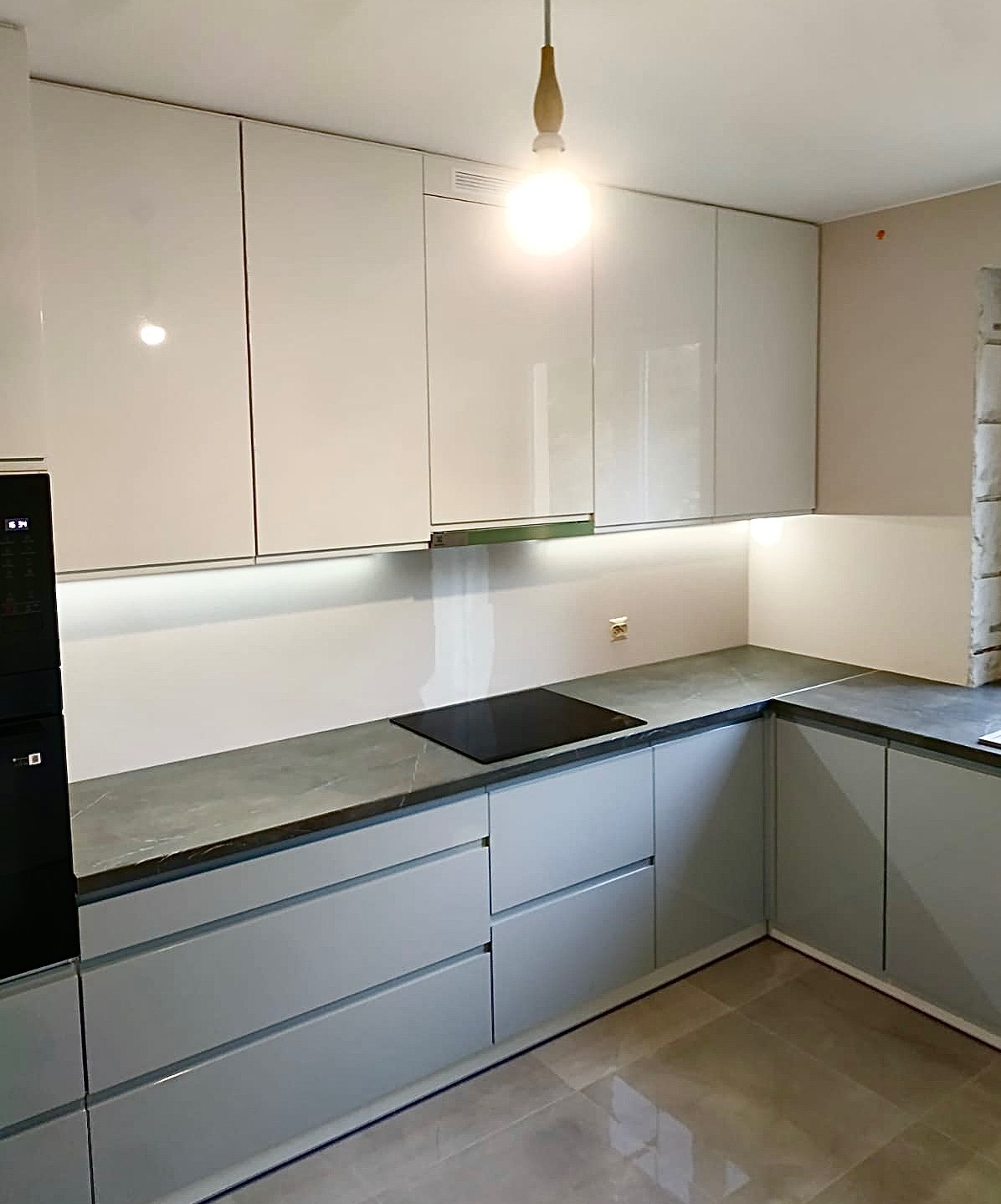Nowoczesna kuchnia z szarymi, lakierowanymi frontami, blatem imitującym kamień i oświetleniem LED pod szafkami wiszącymi. Widoczna płyta indukcyjna i fragment ściany z instalacją.