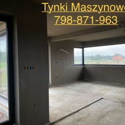 Tynki Kalisz Konin Turek - Surowe wnętrze po tynkowaniu maszynowym: szare ściany, okna z widokiem na zieleń, widoczne instalacje elektryczne. Podłoga betonowa, przygotowana do dalszych prac.