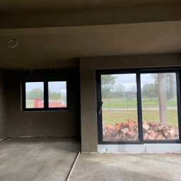 Tynki Kalisz Konin Turek - Wnętrze po nałożeniu tynków: gładkie ściany w odcieniach szarości, widoczne okna z widokiem na zewnątrz, cegły przy oknie balkonowym, surowy stan wykończenia.