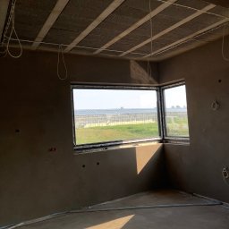 Tynki Kalisz Konin Turek - Pokój po nałożeniu tynku maszynowego, widoczne okna narożne z widokiem na szklarnie. Instalacja elektryczna na suficie. Świeżo otynkowane ściany w odcieniach szarości.