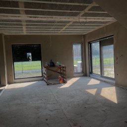 Tynki Kalisz Konin Turek - Surowe wnętrze po tynkowaniu: ściany pokryte szarym tynkiem, widoczne okna z roletami, drewniany stół z narzędziami. Podłoga betonowa, sufit z elementami konstrukcyjnymi.