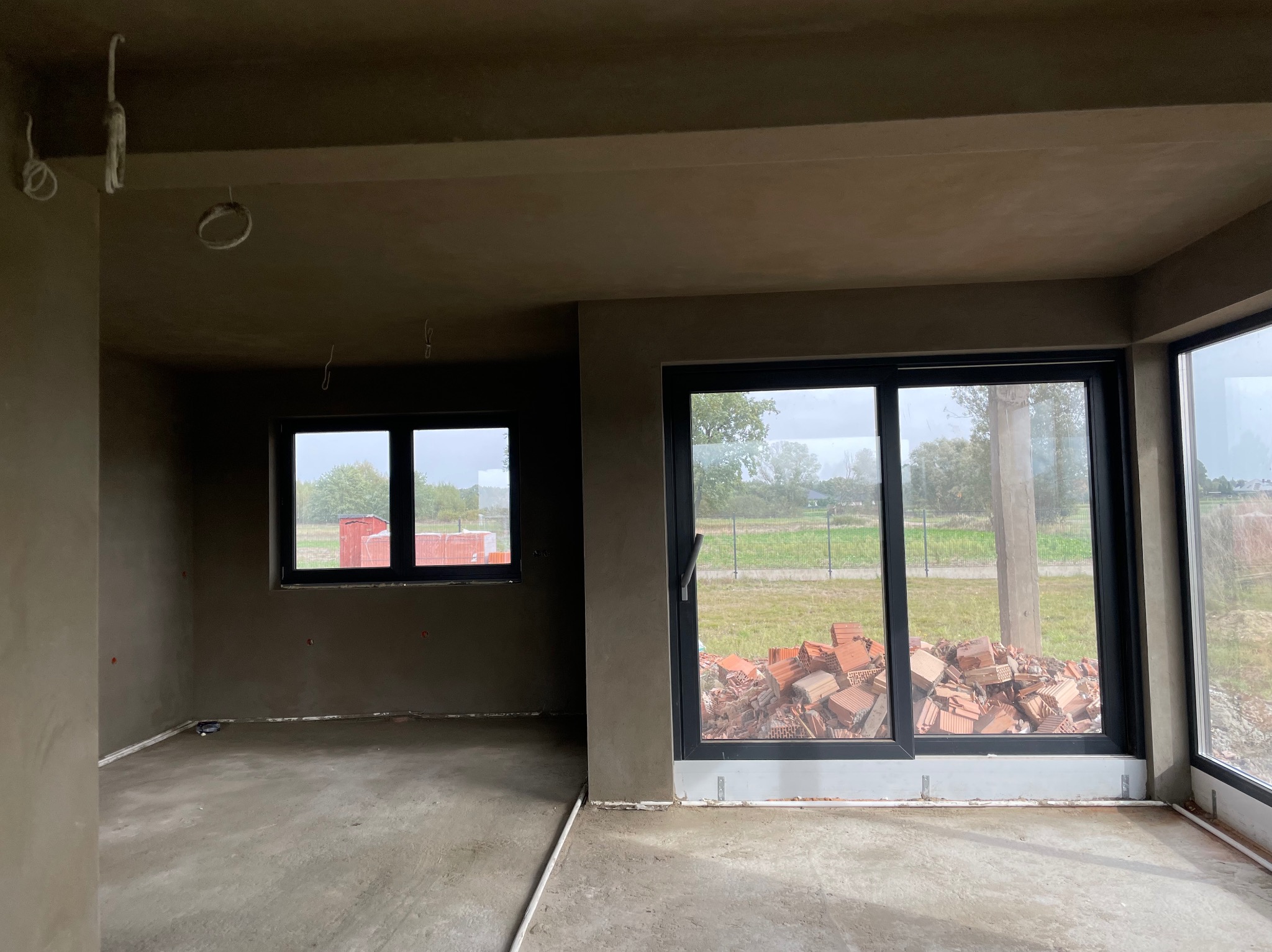 Wnętrze po nałożeniu tynków: gładkie ściany w odcieniach szarości, widoczne okna z widokiem na zewnątrz, cegły przy oknie balkonowym, surowy stan wykończenia.