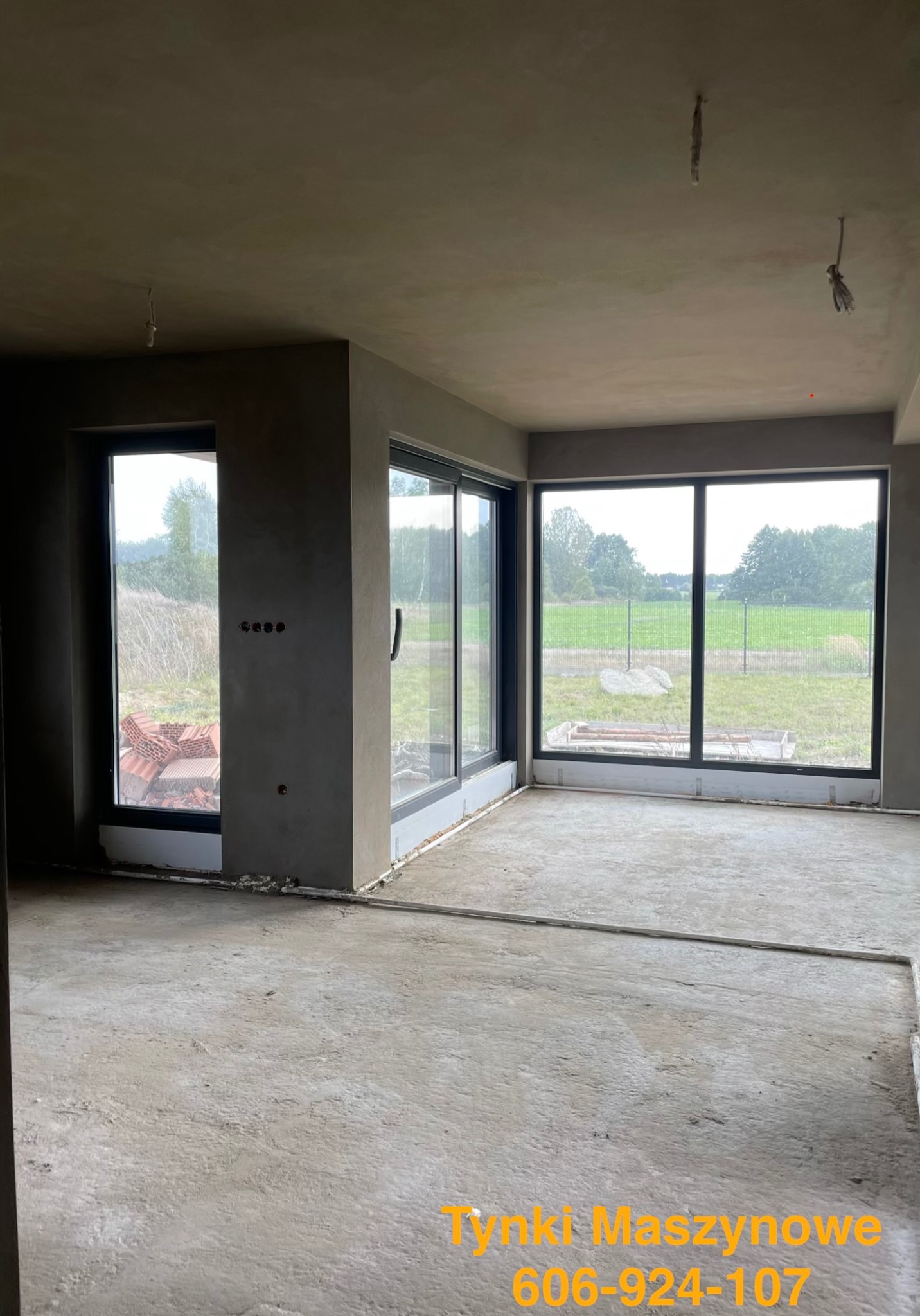 Surowe wnętrze po tynkowaniu maszynowym: szare ściany, duże okna z widokiem na zieleń, betonowa podłoga. Widoczne niedoskonałości i przygotowania do dalszych prac wykończeniowych.
