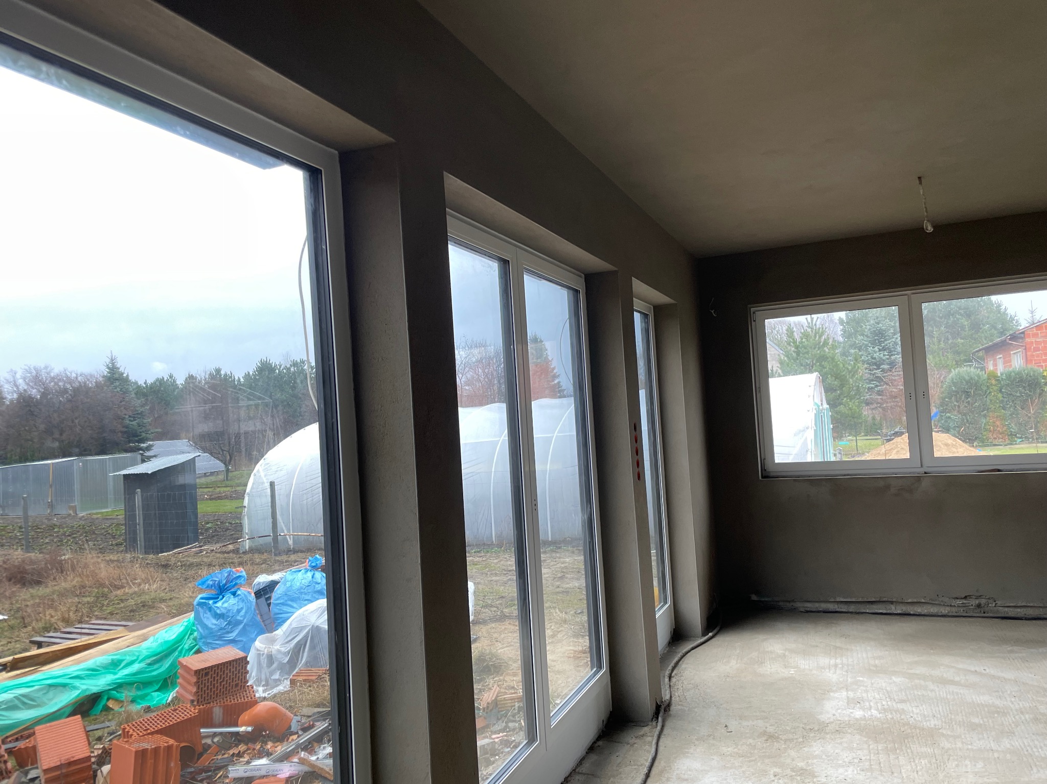 Wnętrze po nałożeniu tynków: gładkie ściany w odcieniach szarości, duże okna z widokiem na zewnątrz, widoczne materiały budowlane. Surowy stan wykończenia.