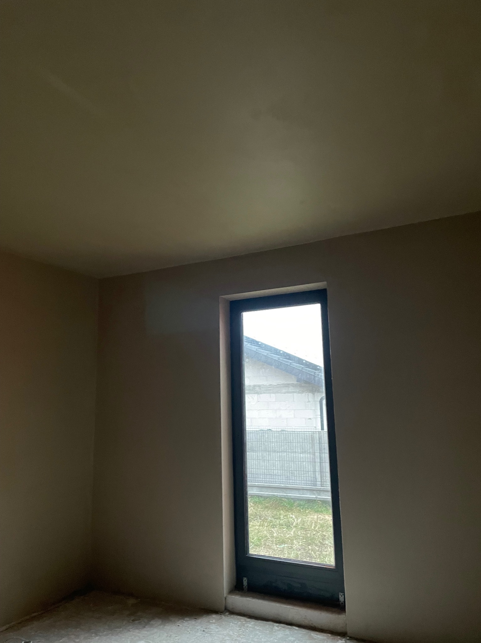 Wnętrze po tynkowaniu maszynowym: gładkie, beżowe ściany w pokoju z oknem. Widoczny fragment podłogi przed wykończeniem i budynek za oknem.