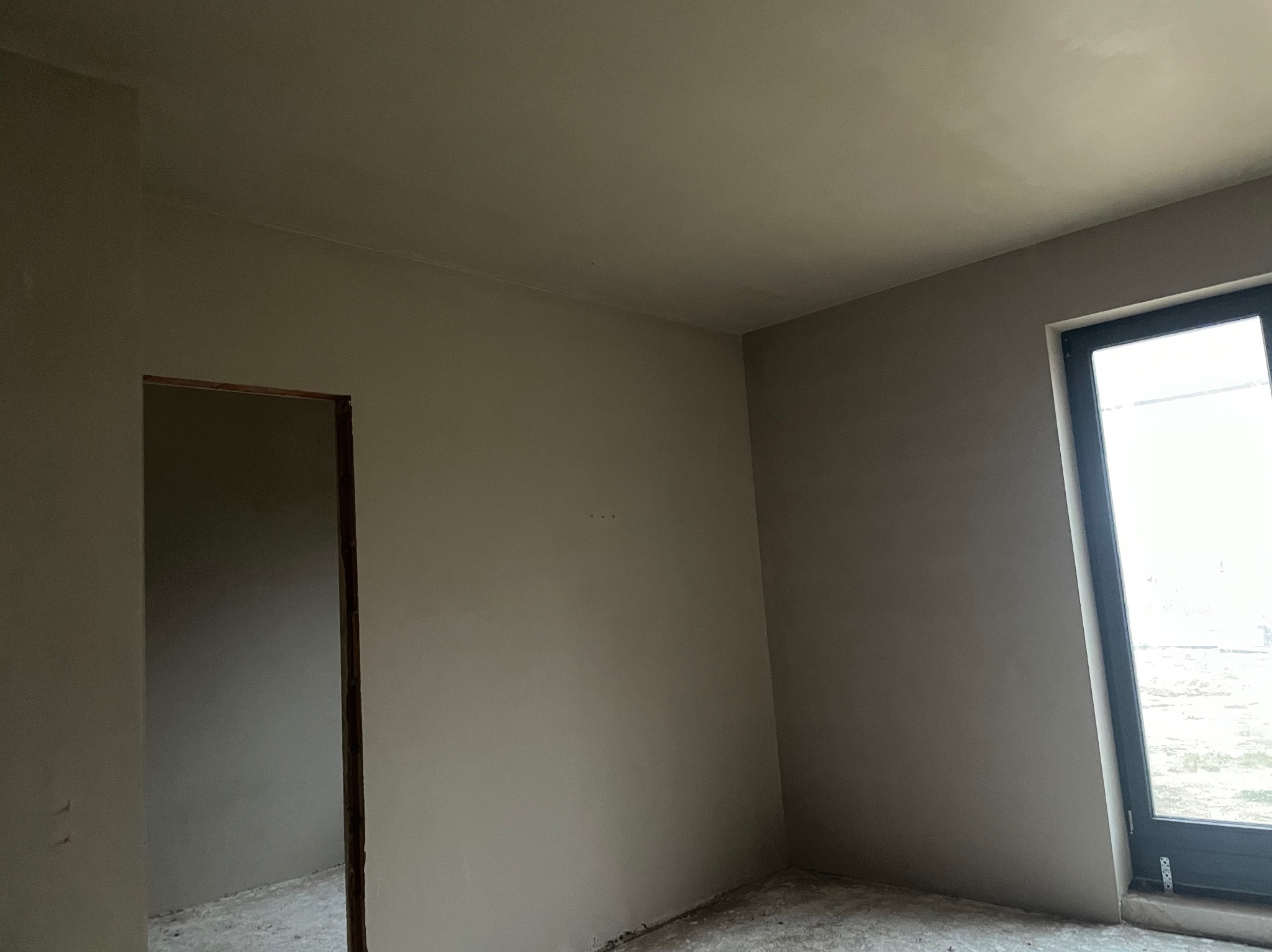 Wnętrze po nałożeniu tynków: gładkie, szare ściany, widoczne surowe wykończenie podłogi, okno z czarną ramą i otwór drzwiowy bez ościeżnicy. Minimalistyczny kadr.