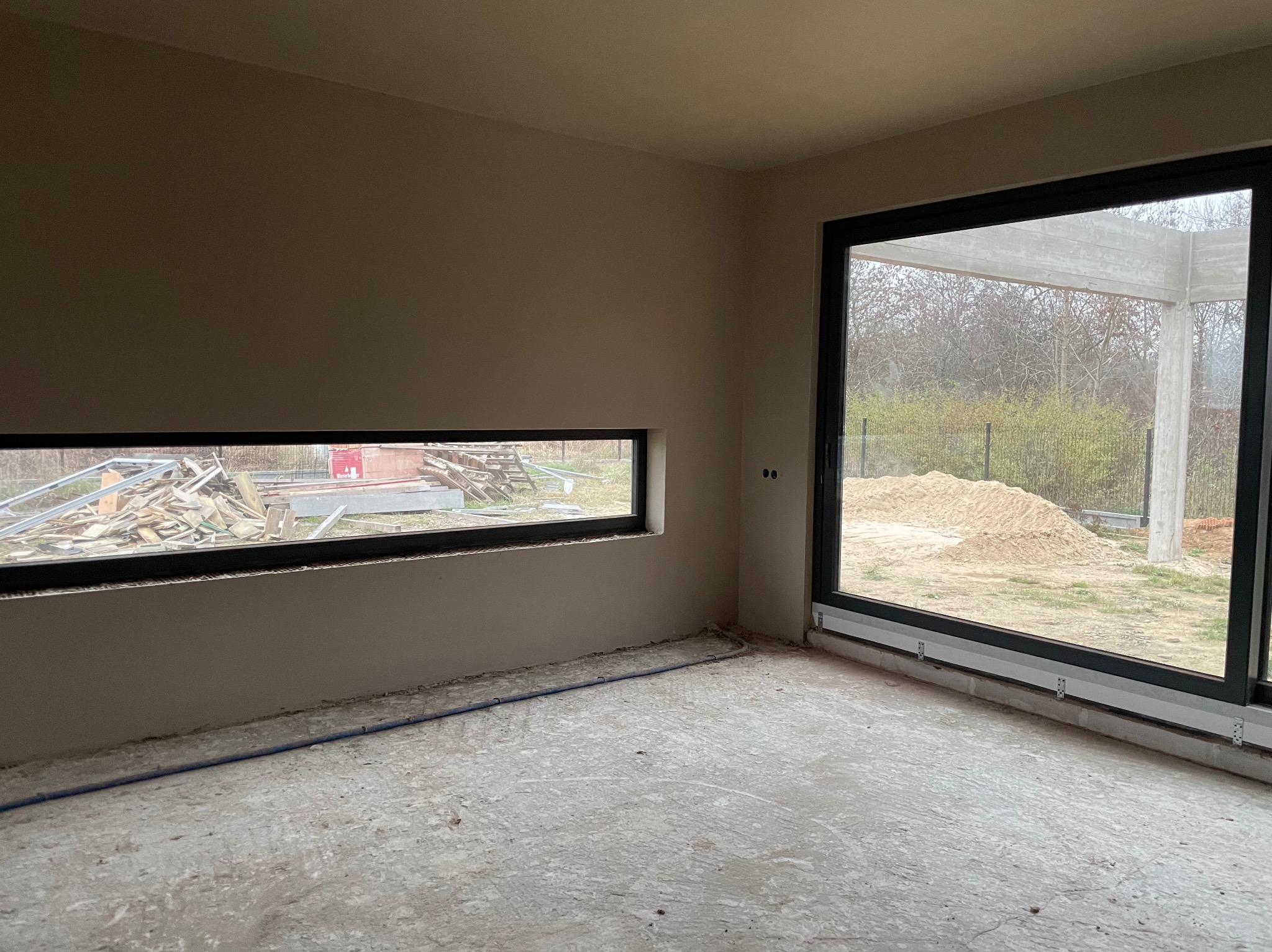Wnętrze po nałożeniu tynków: gładkie, beżowe ściany z oknami, surowa podłoga. Widoczne przygotowania do dalszych prac wykończeniowych. Minimalistyczny, nowoczesny styl.