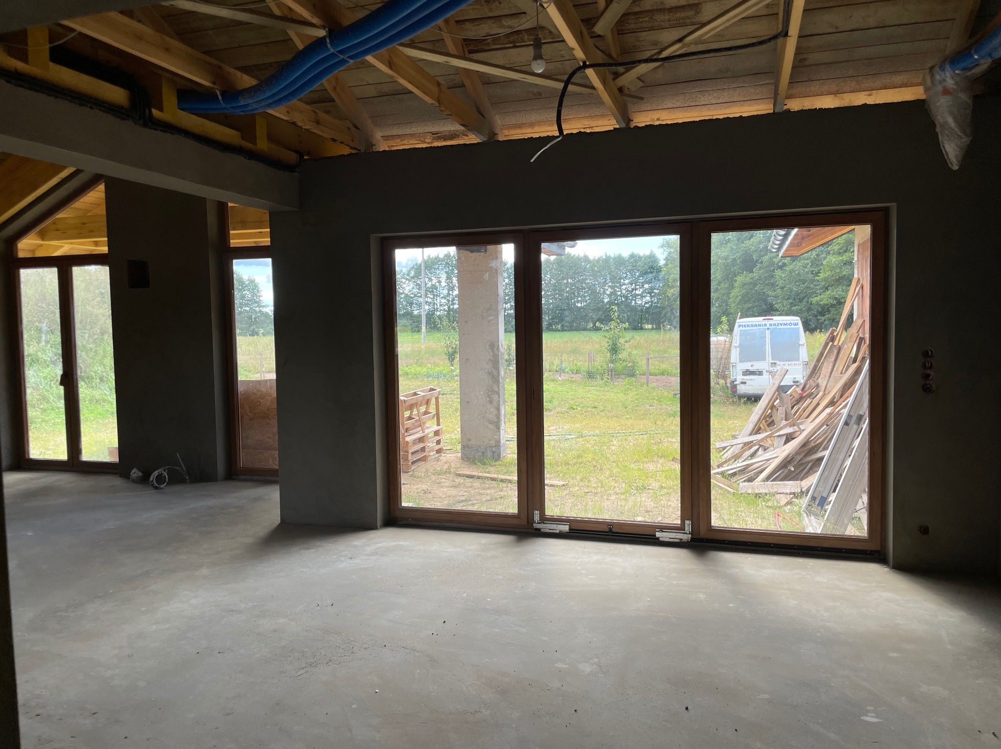 Surowe wnętrze domu po tynkowaniu maszynowym. Widoczne drewniane okna, surowy beton na podłodze i ściany pokryte świeżym tynkiem. Na zewnątrz widok na zieleń.