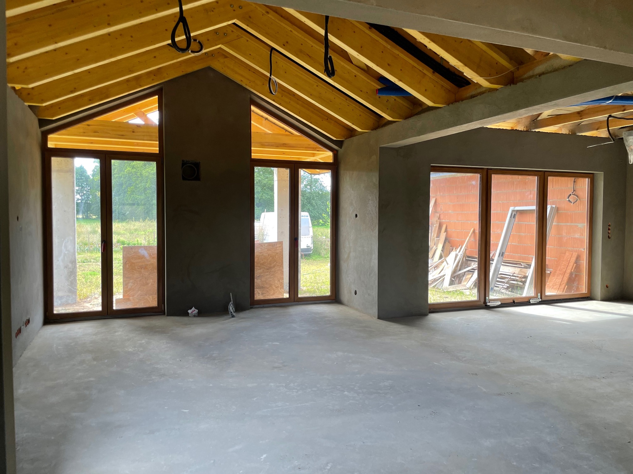 Surowe wnętrze domu w trakcie prac wykończeniowych: szare ściany po tynkowaniu, drewniana więźba dachowa, okna z widokiem na zieleń. Podłoga betonowa.