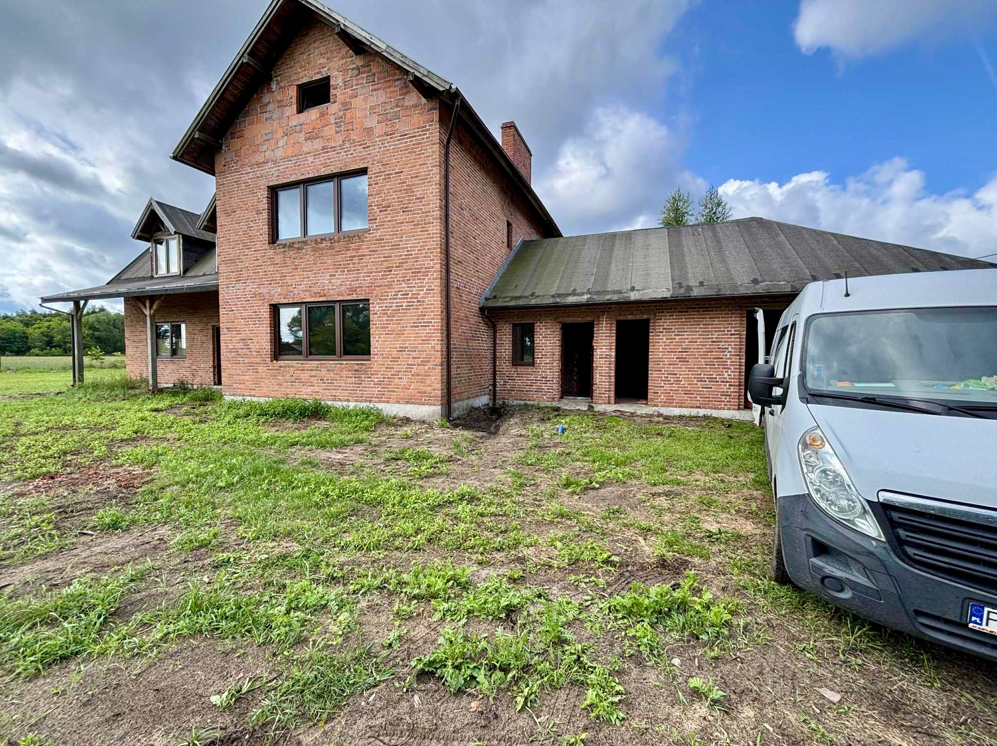 Surowy, dwukondygnacyjny dom z czerwonej cegły w trakcie budowy, z widocznym samochodem dostawczym na tle zachmurzonego nieba. Stan surowy otwarty.