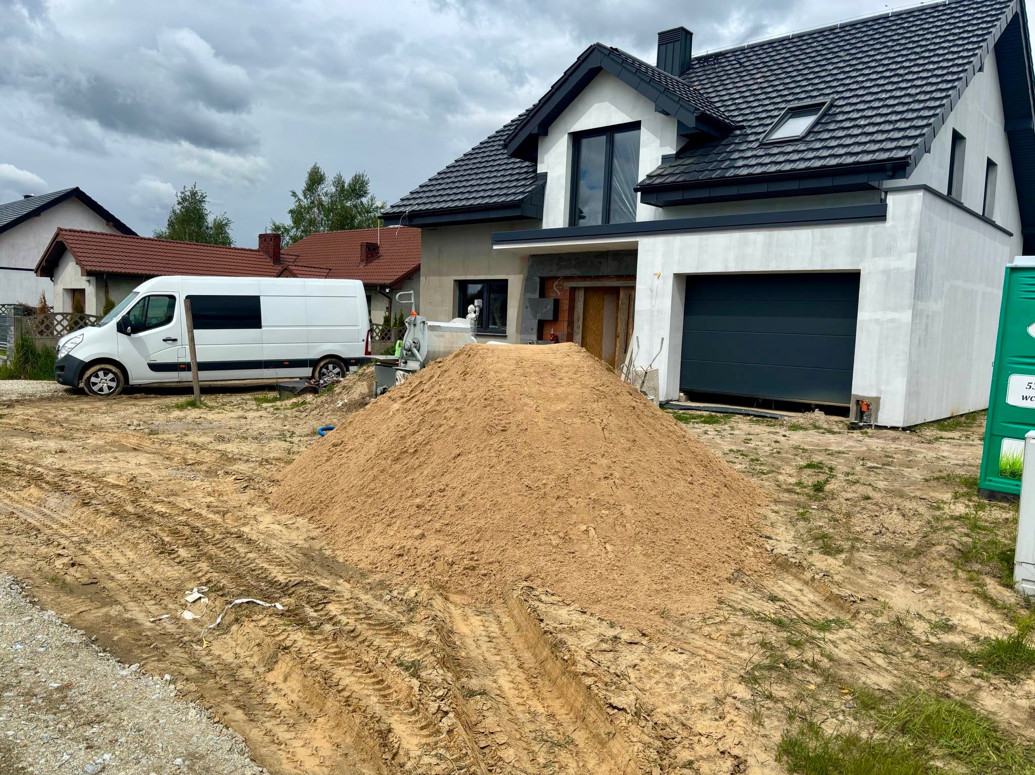 Nowoczesny dom w trakcie tynkowania, z widoczną stertą piasku i samochodem dostawczym na posesji. Szare tynki zewnętrzne i ciemna dachówka.