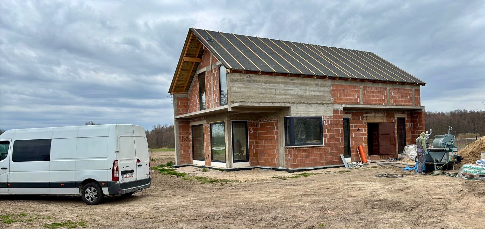 Budowa domu jednorodzinnego w stanie surowym otwartym z widocznym agregatem tynkarskim i samochodem dostawczym na działce budowlanej.