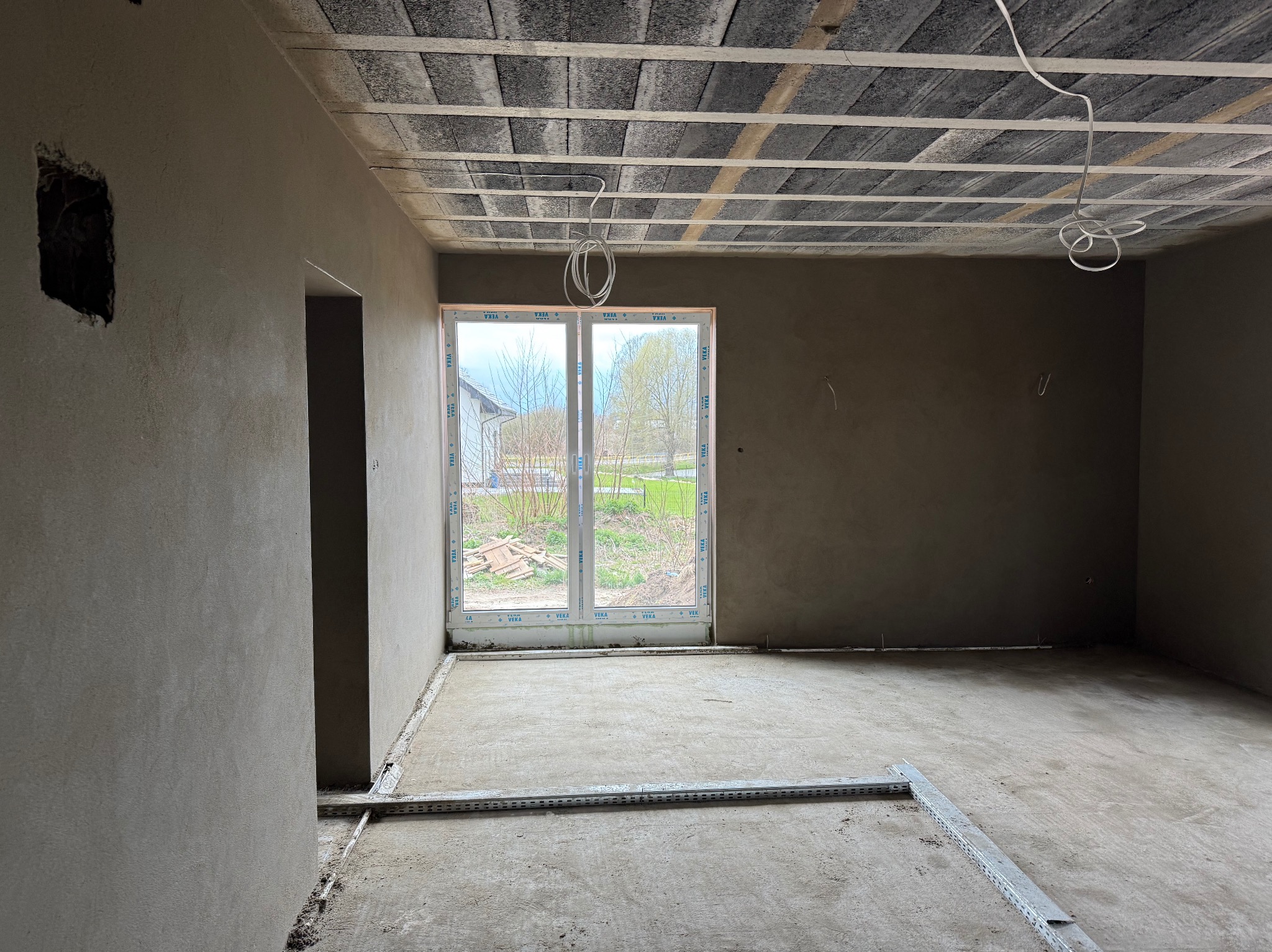 Surowe wnętrze po nałożeniu tynków: ściany pokryte szarym tynkiem, widoczne profile podłogowe, okno z widokiem na zieleń. Przewody zwisają z sufitu.