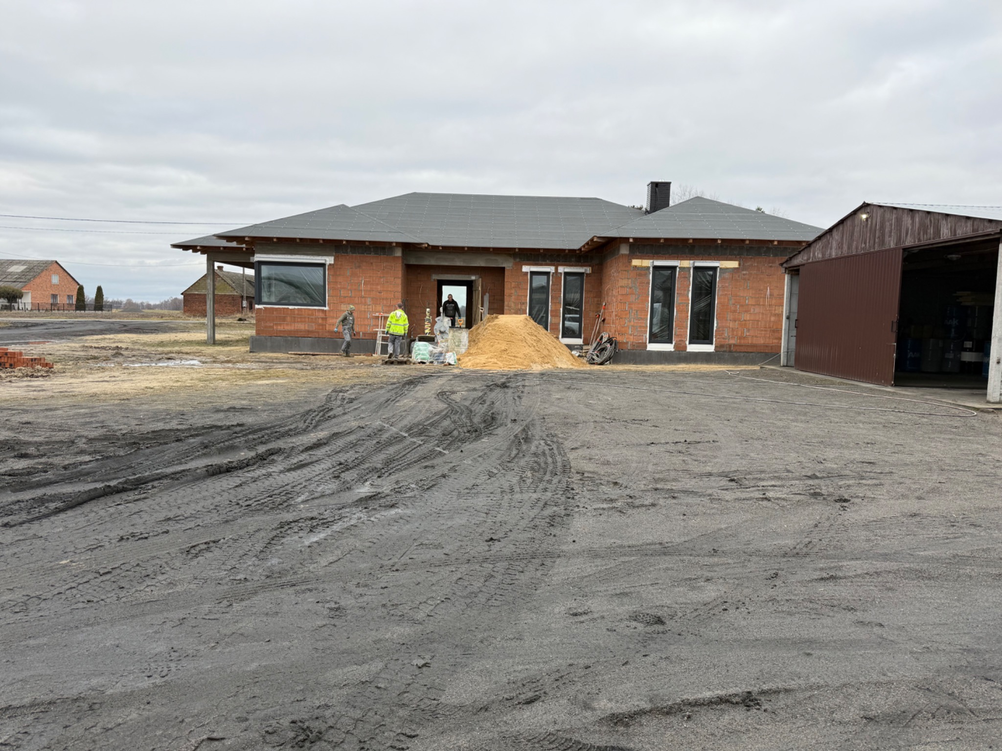 Budowa domu jednorodzinnego w trakcie realizacji: murowane ściany z pustaków, zamontowane okna, przygotowany piasek. Widoczni pracownicy w odblaskowych kamizelkach.